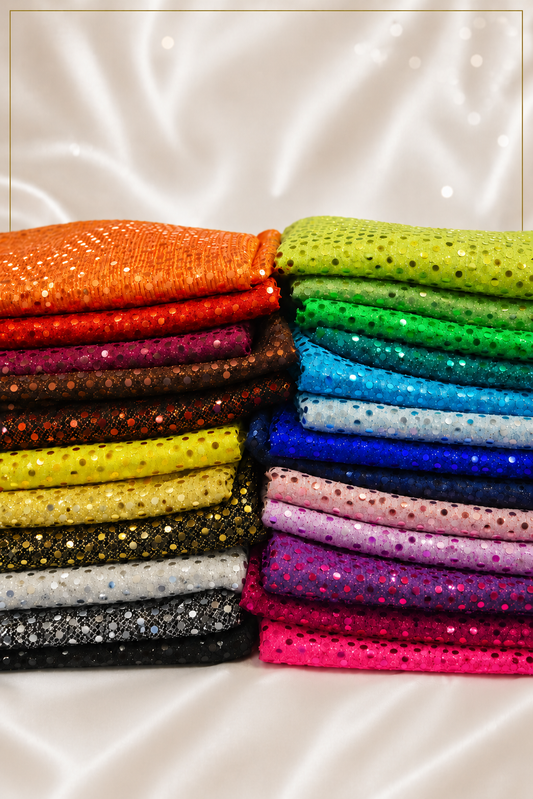 Two stacks of folded sequined fabrics in a rainbow of colors, including orange, red, pink, yellow, green, blue, purple, and black, arranged on a white satin background.