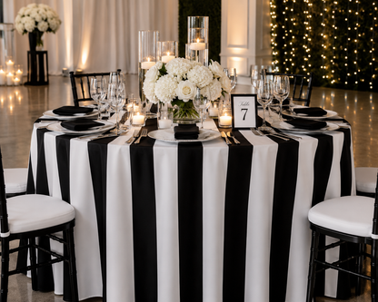 A round event table set with a bold black and white vertically striped tablecloth, white floral centerpieces, clear glass candleholders, wine glasses, black plates, and white chair covers in a warmly lit indoor venue.
