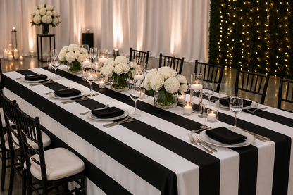 A long banquet table set for guests with a bold black and white striped tablecloth, white floral centerpieces, black napkins, white plates, and black wooden chairs in an indoor event space with warm lighting and string lights.