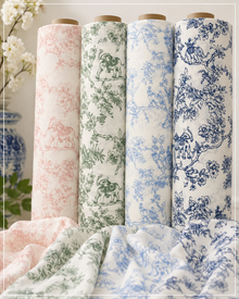 Four fabric rolls with wooden cores stand upright, each printed with a classic toile pattern in a different color: soft pink, sage green, light blue, and dark navy blue. A matching folded fabric is draped in the foreground.