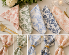 Six folded fabric napkins with floral toile patterns are displayed on a table setting, shown in coral pink, sage green, light blue, navy blue, and peach. The top row shows the napkins styled with gold-toned flatware and a candle, while the bottom row is a vertical strip of close-up detail views of each napkin's fabric pattern and texture.