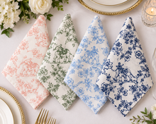 Four folded cloth napkins with classic toile patterns are arranged on a white tablecloth. The napkins feature different colorways: pink, sage green, light blue, and navy blue.