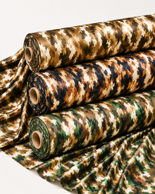 Three rolls of camouflage fabric stacked diagonally, with additional fabric draped out in front, showing a multi-colored camo pattern in brown, tan, green, and black on a light gray background.