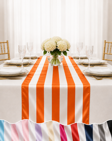 A white tablecloth with bold orange vertical stripes and a ruffled hem, styled on a dining table with white plates, wine glasses, a floral centerpiece, and gold chairs.