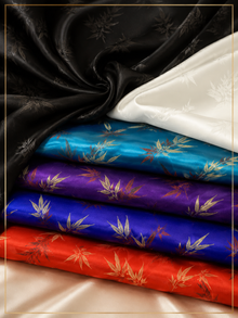 Four folded satin fabrics in black, white, turquoise blue, purple, and red, each printed with a gold-toned bamboo leaf pattern, arranged on a beige satin surface.