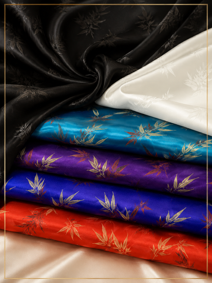 Four folded satin fabrics in black, white, turquoise blue, purple, and red, each printed with a gold-toned bamboo leaf pattern, arranged on a beige satin surface.