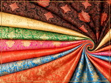 A collection of richly colored brocade fabrics arranged in a spiral fan, showcasing vibrant shades of red, blue, pink, green, gold, and brown with intricate metallic floral patterns.