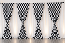 Five black and white checkered curtains tied at the bottom, hanging in front of sheer white drapes on a rod, with a reflective dark floor below.